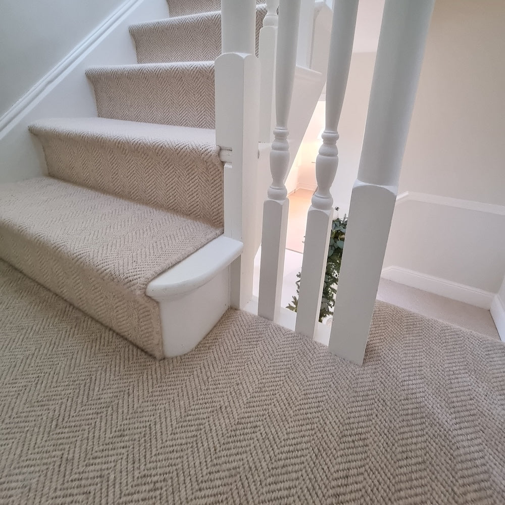 A beige herringbone-patterned carpet installed on a staircase and landing, with white wooden balusters and steps also covered in matching carpet.