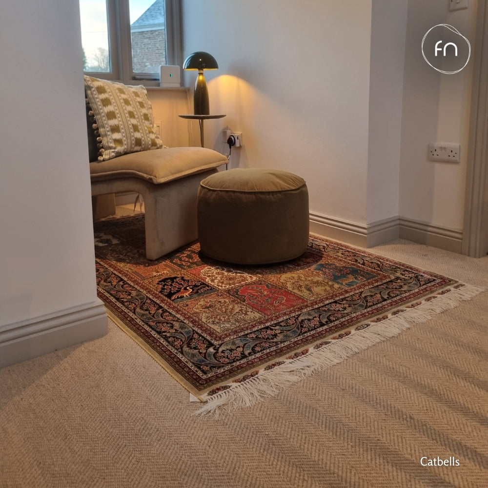 A styled interior corner showing a patterned area rug on a light herringbone wool carpet, with a beige upholstered armchair, a round brown ottoman, a small side table, and a black dome lamp.