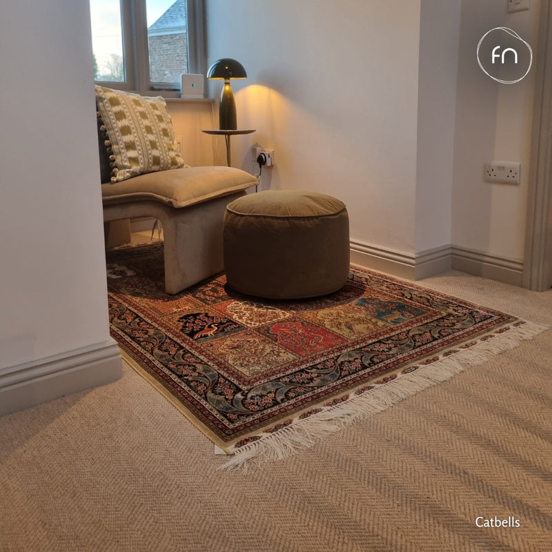 A styled interior corner showing a patterned area rug on a light herringbone wool carpet, with a beige upholstered armchair, a round brown ottoman, a small side table, and a black dome lamp.