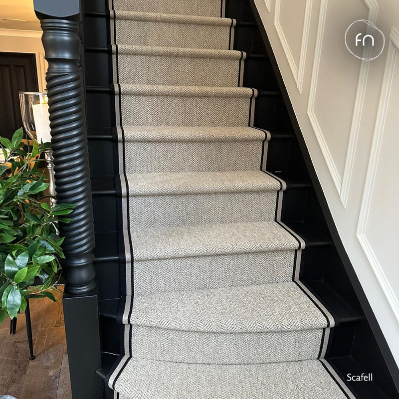A staircase with carpeted steps in a light grey herringbone pattern, bordered by black trim, set against a white panelled wall with a dark wooden banister visible on the left.