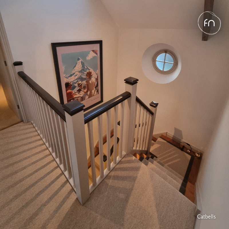 A staircase & landing carpeted in a light beige wool herringbone with a white spindle railing and dark wooden handrails, featuring a framed mountain artwork on the wall and a round window high up in the stairwell.
