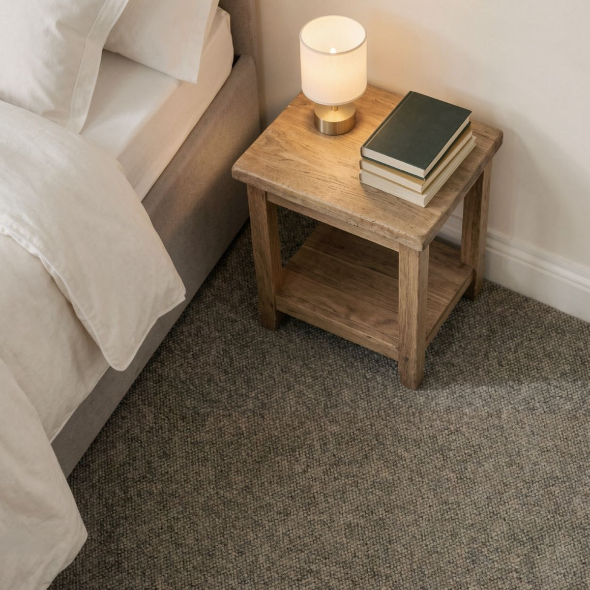 A rustic wooden nightstand with an open shelf sits beside a bed in a bedroom. A lit table lamp and a stack of three books rest on the nightstand, which is placed on a textured gray carpet against a white wall.