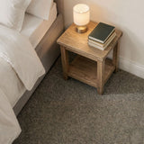 A rustic wooden nightstand with an open shelf sits beside a bed in a bedroom. A lit table lamp and a stack of three books rest on the nightstand, which is placed on a textured gray carpet against a white wall.