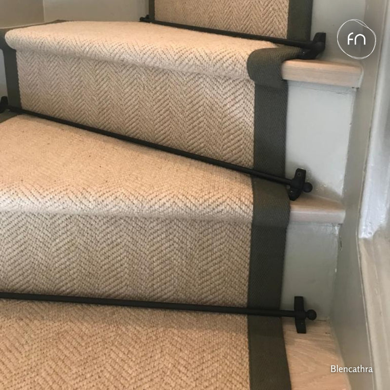A close-up of carpeted stairs with an undyed wool herringbone pattern, dark grey side borders, and black metal stair rails.
