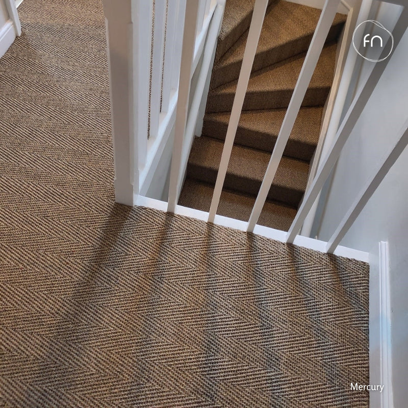 A close-up view of a sisal carpeted staircase landing with a herringbone pattern in brown, beige, and grey tones, bordered by white baseboards and white stair railings.