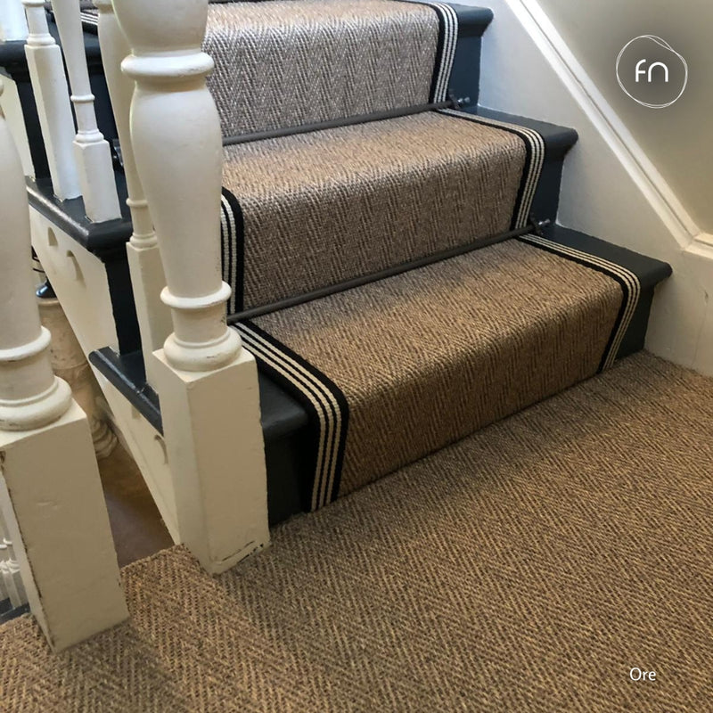A set of steps carpeted with a brown and beige herringbone pattern sisal, bordered by black and white stripes, installed in a home interior with white wooden banisters.